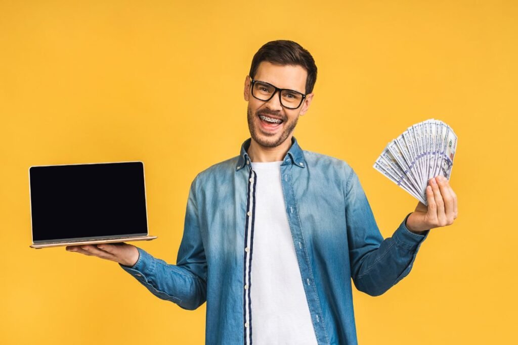 happy man with laptop in one hand and cash on other in front of yellow background