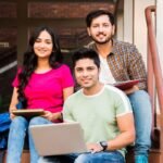 Three students sitting on college steps, one with a laptop and two with books