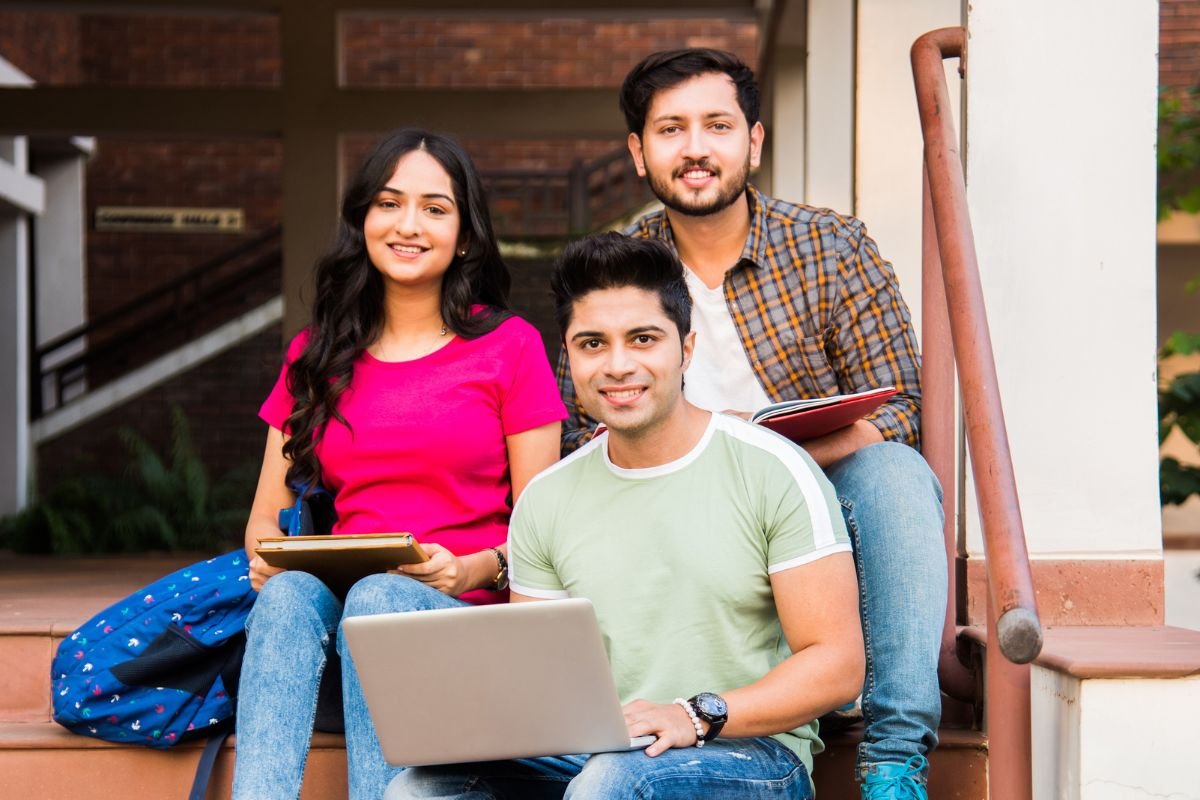 Three students, one girl in a pink top and two boys in shirts, are sitting on a ladder of a college, one holding a laptop and two holding books.