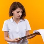 young girl with short hair wearing polos shirt holding clipboard with document pages looking it with serious face
